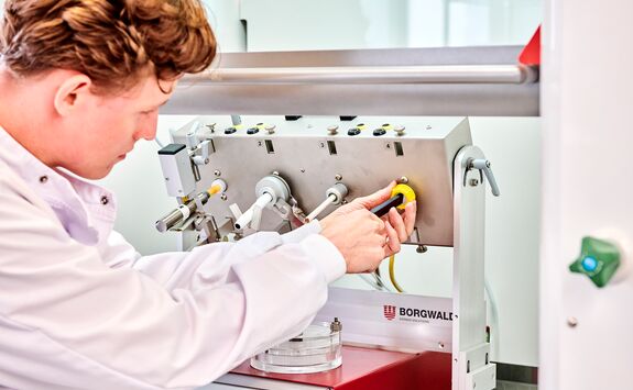 Person in a white lab coat adjusting a yellow knob on a machine. The machine has various buttons, levers, and controls.